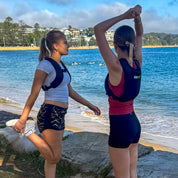 Two women stretching on a rocky beach with a scenic view of water and buildings in the background.